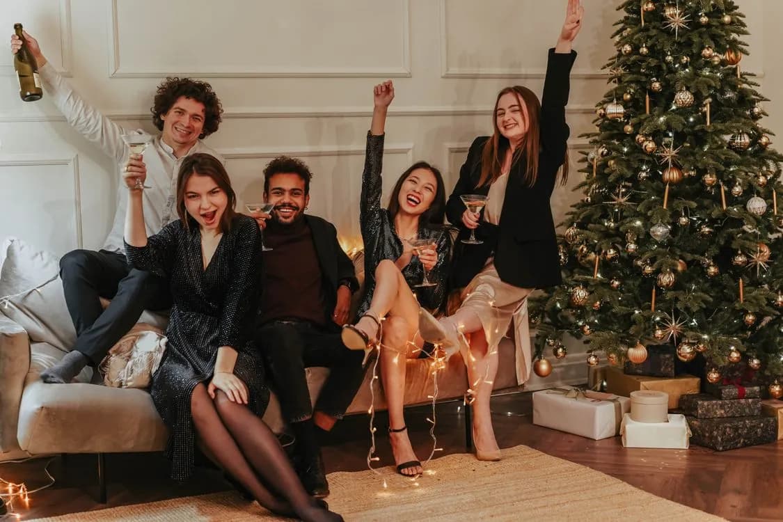 Friends celebrating with drinks beside a decorated Christmas tree in a living room
